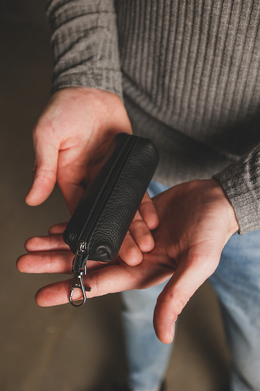 A close-up of a person holding a small black textured leather zippered pouch with a silver metal clip in their hands.