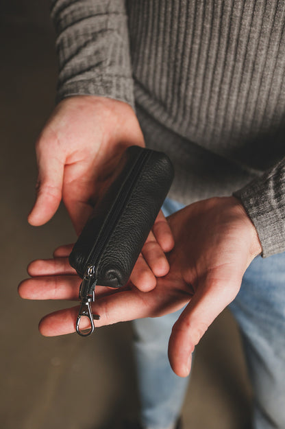 A close-up of a person holding a small black textured leather zippered pouch with a silver metal clip in their hands.