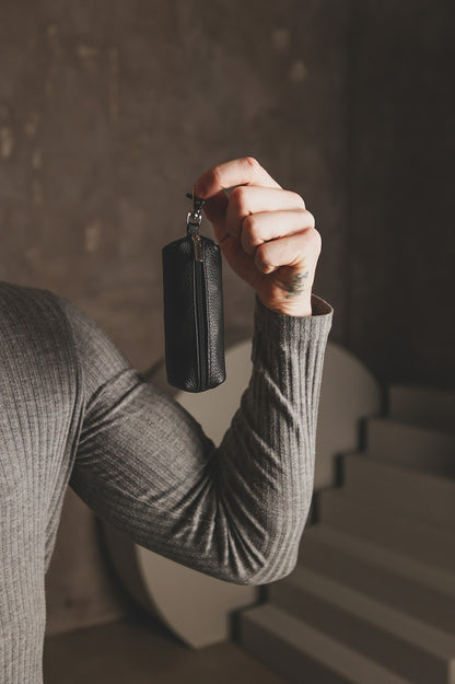 A person in a ribbed gray long-sleeve top holds up a small black textured leather key pouch with a silver zipper and keyring, set against a moody, dimly lit background with concrete walls and stairs.