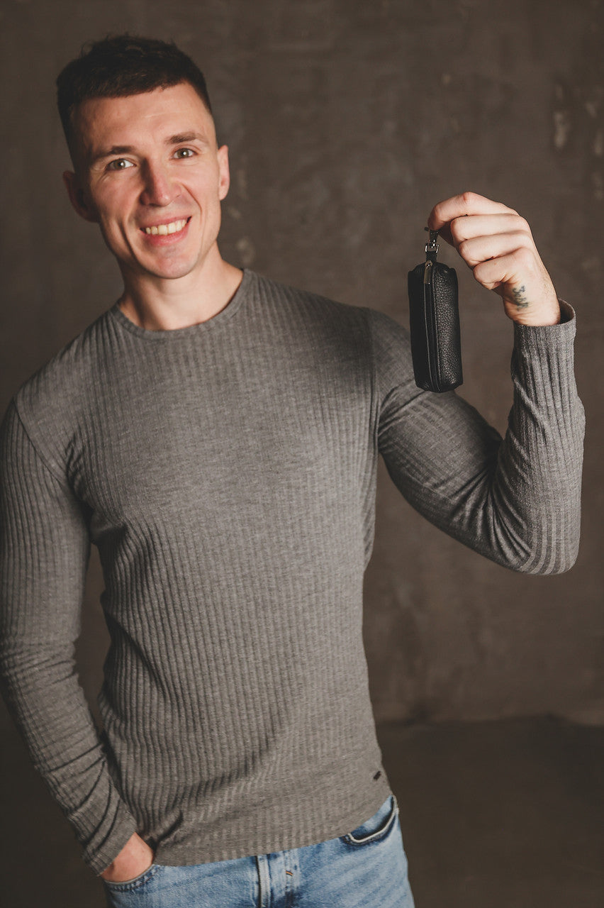 A smiling man wearing a gray ribbed long-sleeve sweater and blue jeans holds up a small black leather pouch or wallet in his raised hand against a textured brown studio background.