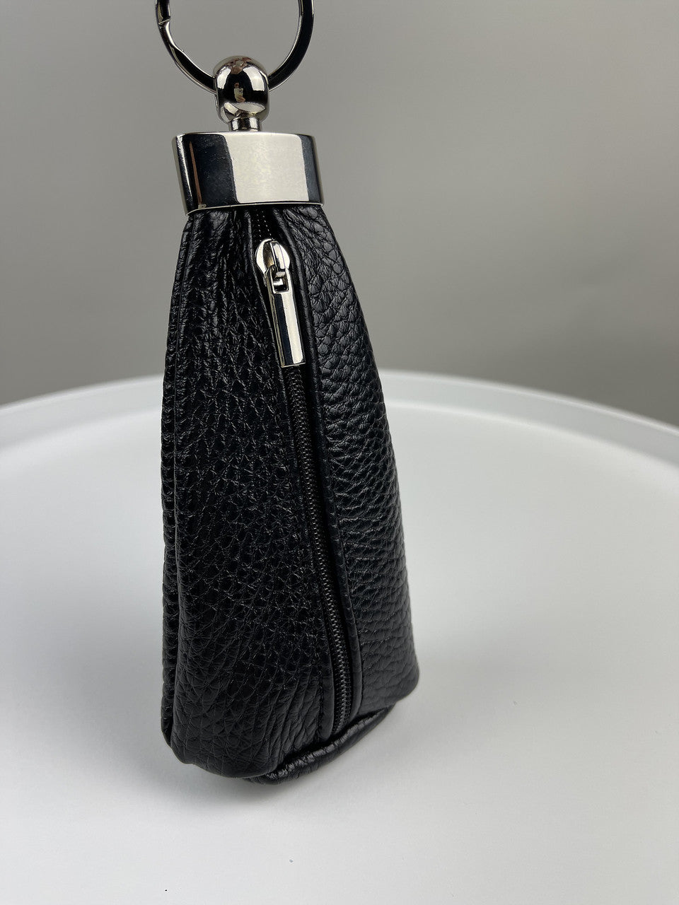 A black pebbled leather key pouch with a silver-tone metal clasp and keyring, standing upright on a white surface against a gray background.