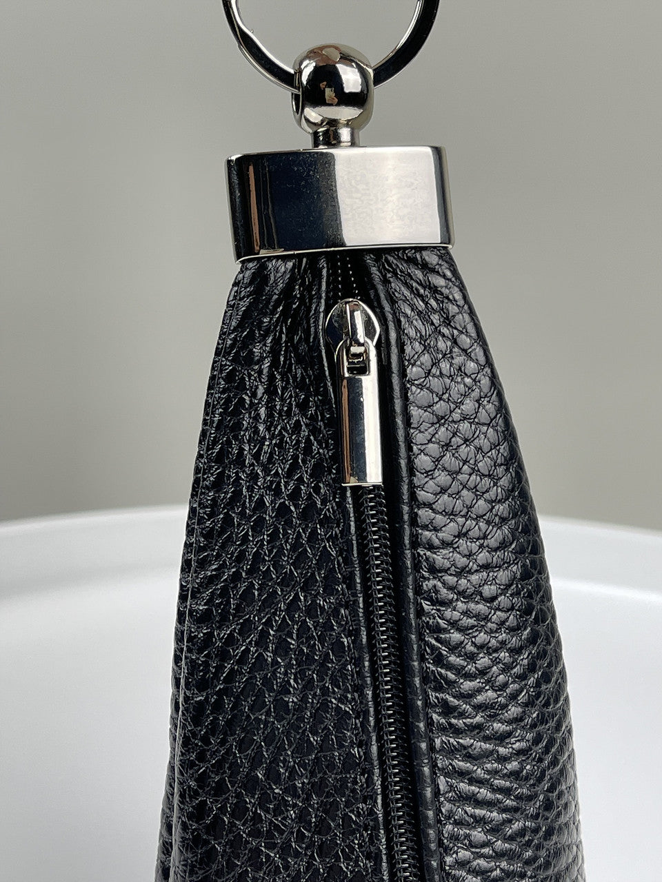 Close-up of a black pebbled leather zippered pouch or clutch with a polished silver-tone metal ring and bar clasp, shown against a neutral gray background.