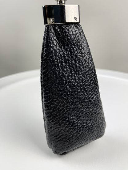 A close-up of a black pebbled leather accessory, likely a small pouch, case, or strap component, with a silver-toned metal cap, standing upright on a white surface against a light gray background.