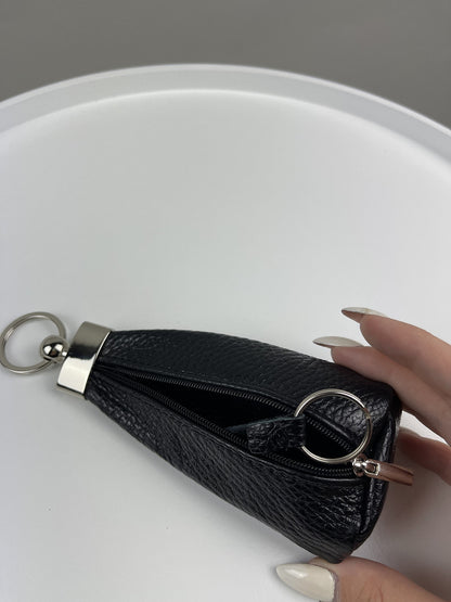 A small black textured leather zip pouch with silver-tone hardware, held by a hand with long nude manicured nails against a white circular surface.