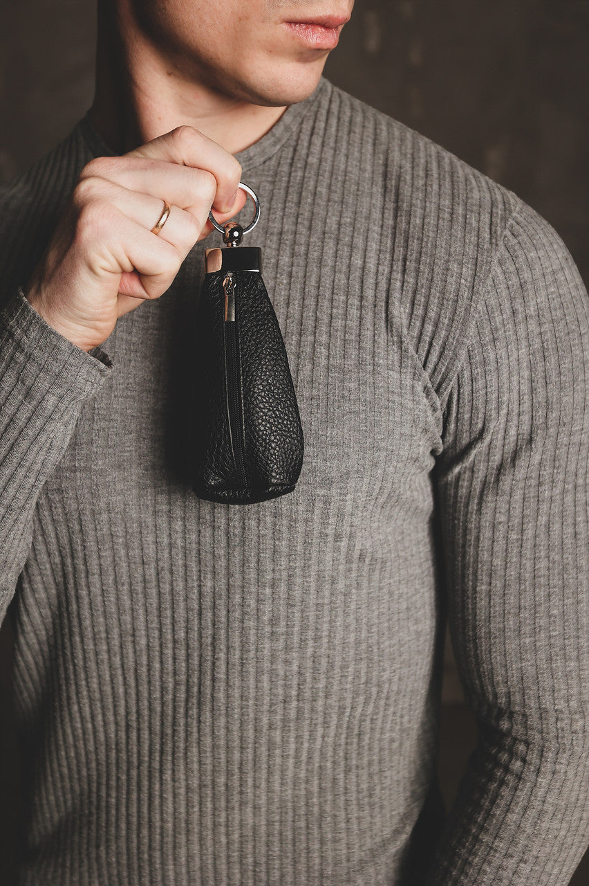 A close-up of a man wearing a gray ribbed knit sweater, holding a small black textured leather pouch with a key ring and gold-tone zipper pull.