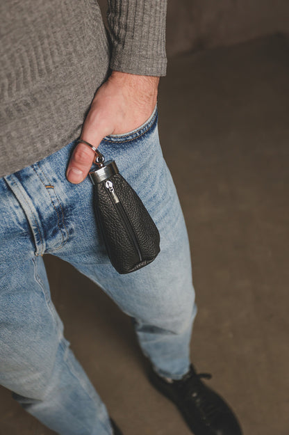 A close-up lifestyle shot of a person wearing light blue jeans and a gray ribbed sweater, holding a small black textured leather bottle pouch with a silver zipper.