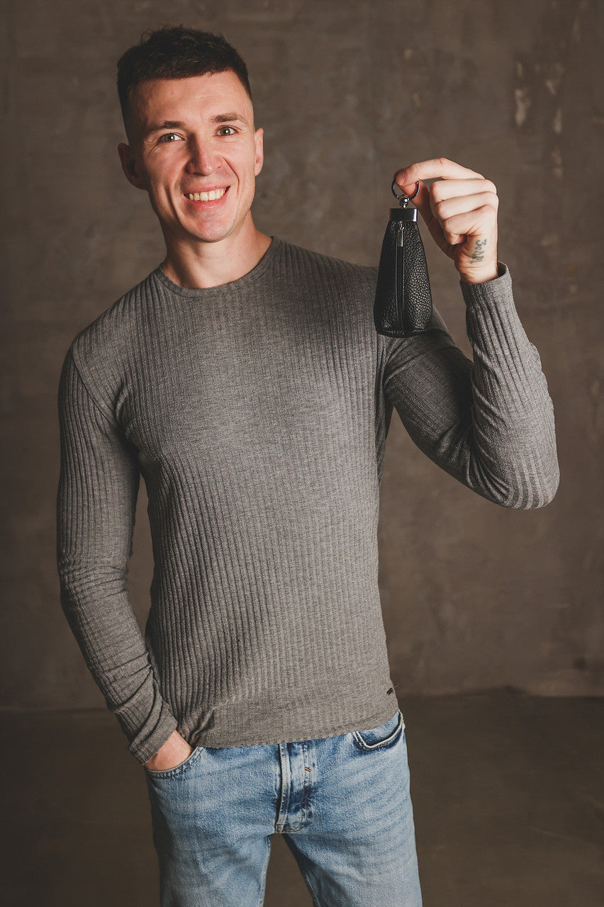 A smiling man wearing a gray ribbed long-sleeve sweater and blue jeans holds up a small black leather pouch or wallet attached to a key ring against a textured gray studio background.