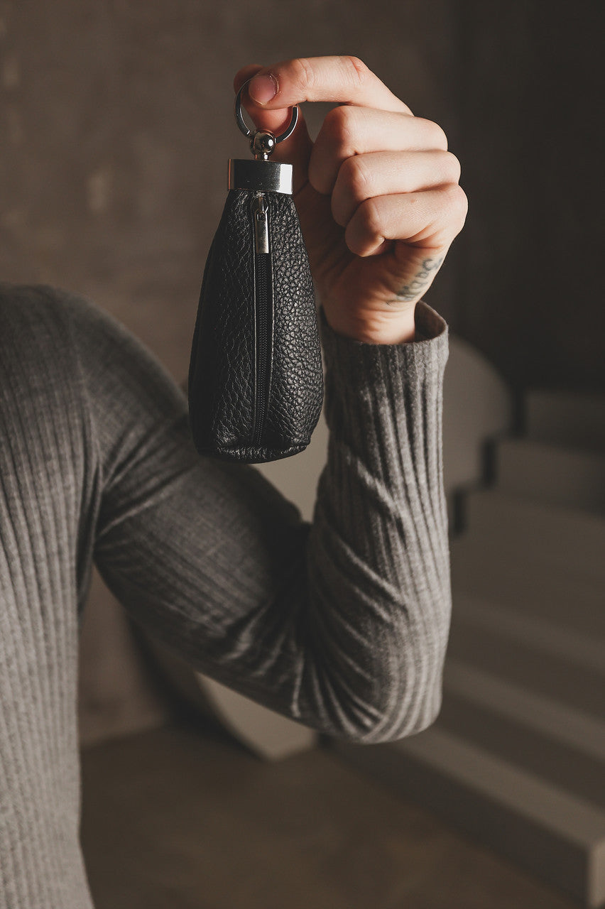 A person in a ribbed gray long-sleeve top holds up a small black pebbled leather keychain bag with a silver metal ring and zipper pull in a dimly lit interior setting.
