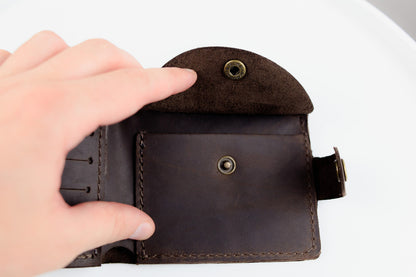 A close-up of a dark brown leather wallet being held open by a hand, showing the interior card slots, snap button, and strap closure against a white background.