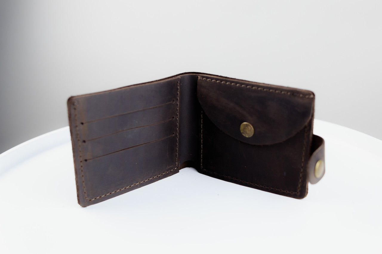 A dark brown leather bifold wallet shown open, displaying its interior card slots and flap pocket with brass-tone snap buttons, set on a white surface against a light gray background.