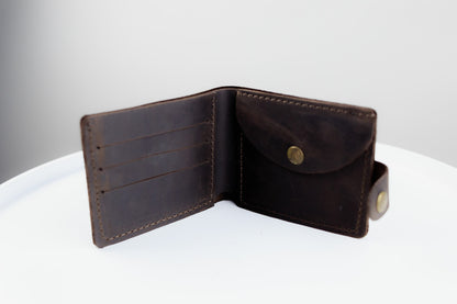 A dark brown leather bifold wallet shown open, displaying its interior card slots and flap pocket with brass-tone snap buttons, set on a white surface against a light gray background.