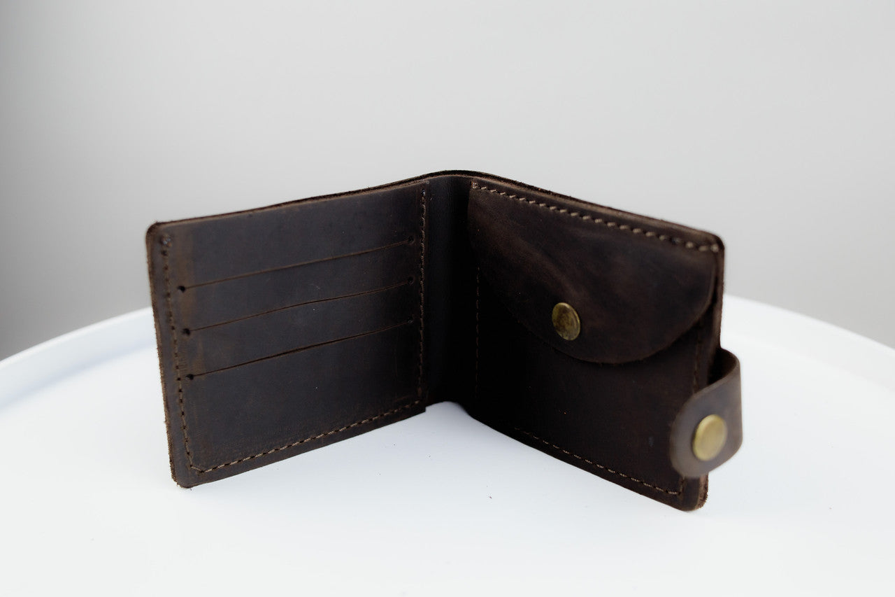 A dark brown leather bi-fold wallet shown open, displaying its interior card slots and pocket, set against a clean white and light gray background.