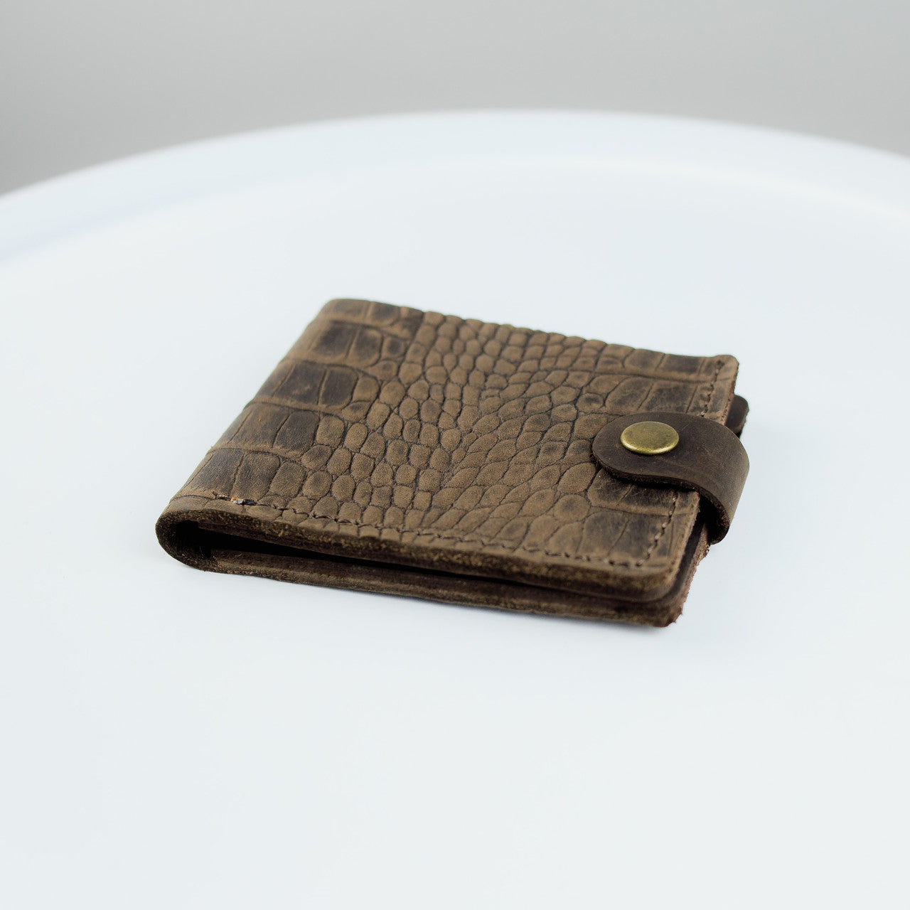 A brown textured leather wallet with a crocodile pattern and a brass snap button closure, resting on a white surface.