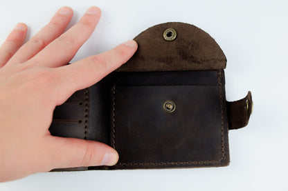 A close-up of a dark brown leather wallet being held open by a hand, showing the interior card slots, snap-button closure, and strap detail against a plain white background.