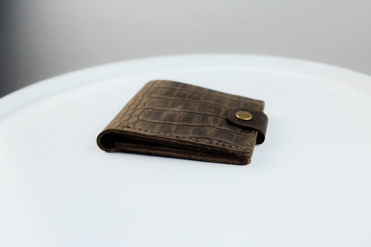 A brown leather bifold wallet with an embossed crocodile pattern and a brass snap button, resting on a white round surface.