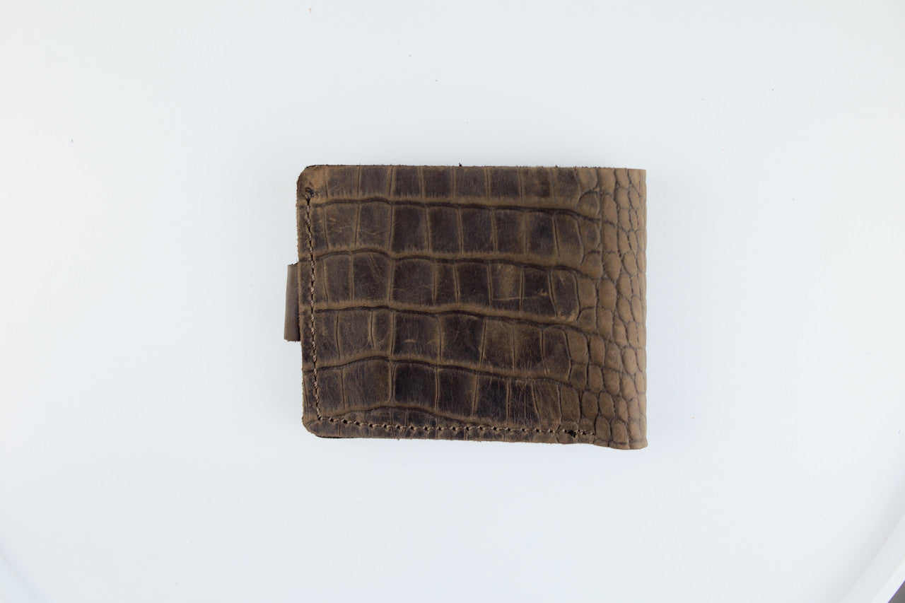 A brown leather bifold wallet with an alligator or crocodile-embossed texture, shown closed on a plain white background.
