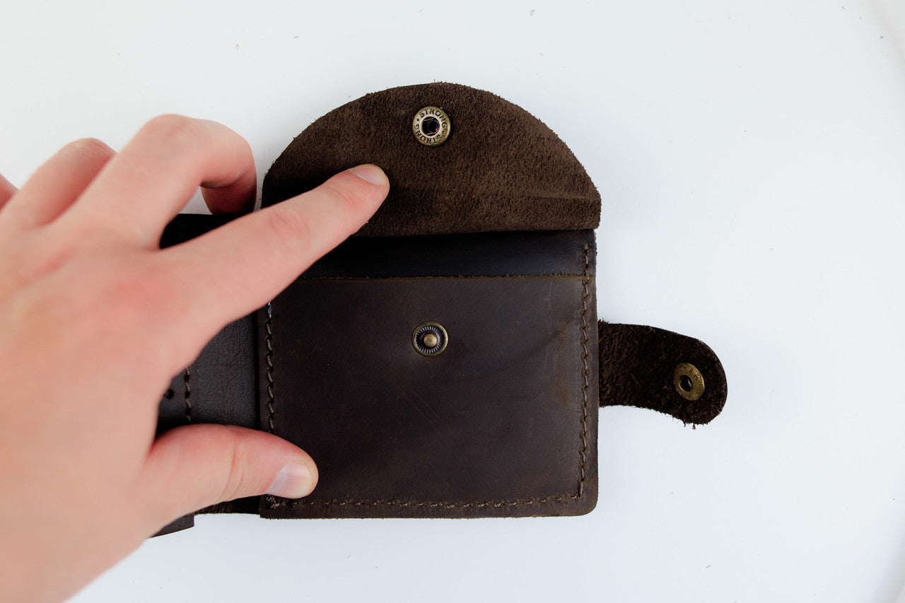A dark brown leather card holder wallet with a suede flap and brass snap buttons, held open by a hand against a plain white background.