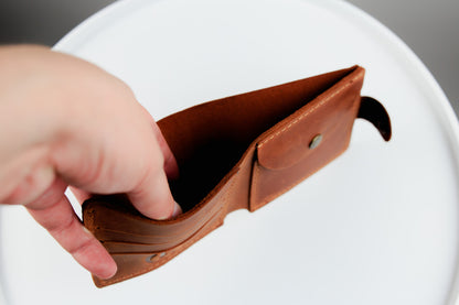 A hand holding a brown leather card holder or wallet with a small metal snap button, shown against a white circular background.