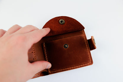 A close-up of a person's hand holding open a small brown leather card holder or wallet with metal snap buttons, set against a plain white background.