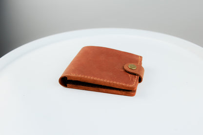 A brown leather bi-fold wallet with a snap button closure, resting on a white circular surface against a neutral gray background.