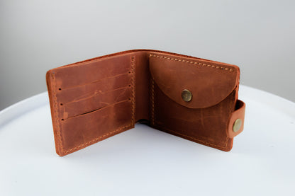 A brown leather bi-fold wallet shown open, displaying its interior card slots and a front pocket with a snap button closure, set against a neutral gray and white background.