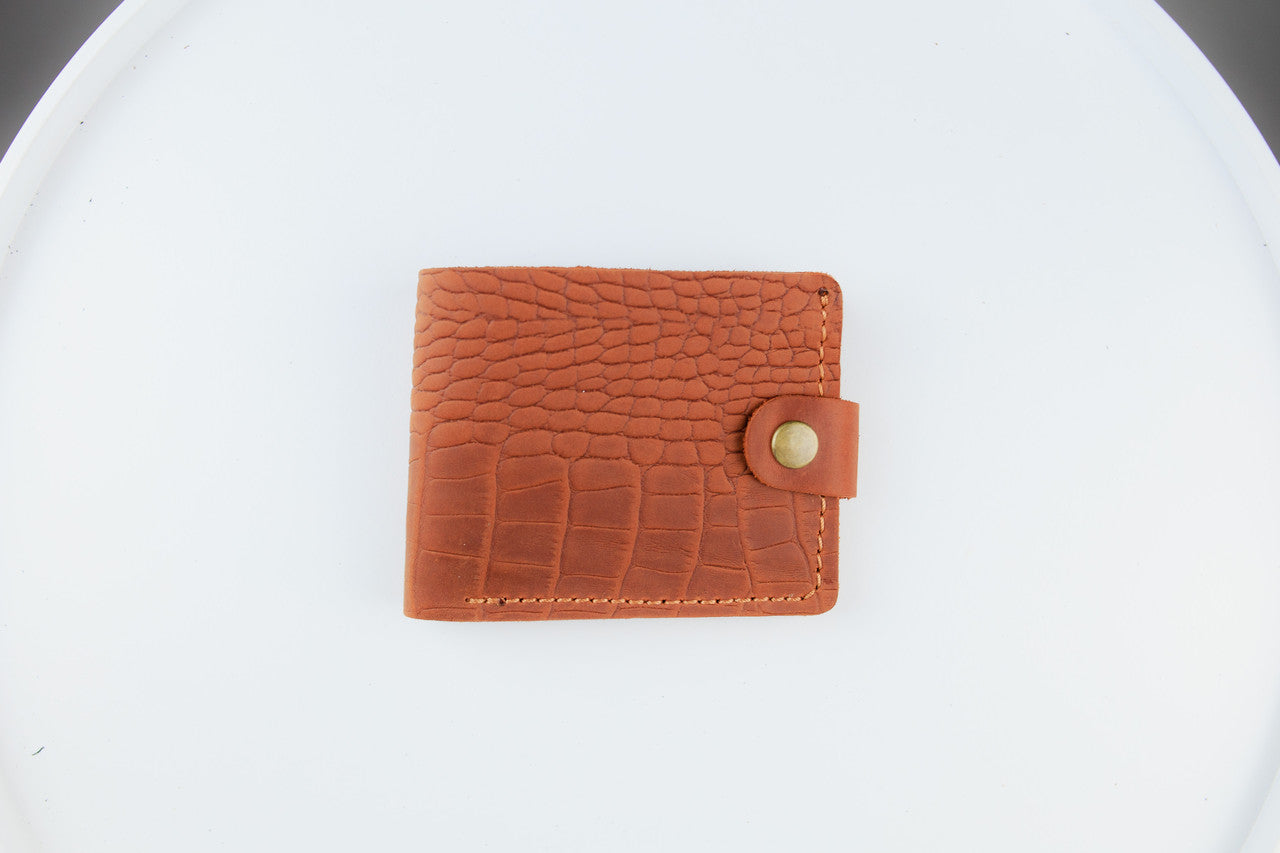 A brown crocodile-embossed leather bifold wallet with a snap-button closure, displayed on a clean white background.