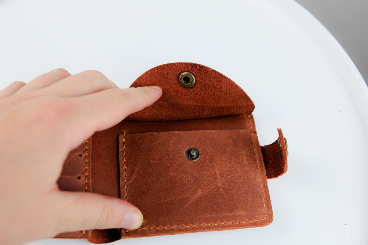 A close-up of a person's hand holding open a small brown leather wallet or card holder, showing the interior compartments and metal snap closures against a white background.