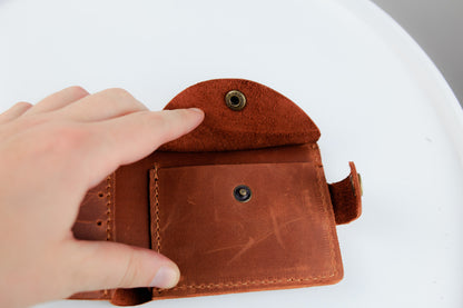 A close-up of a person's hand holding open a small brown leather wallet or card holder, showing the interior compartments and metal snap closures against a white background.