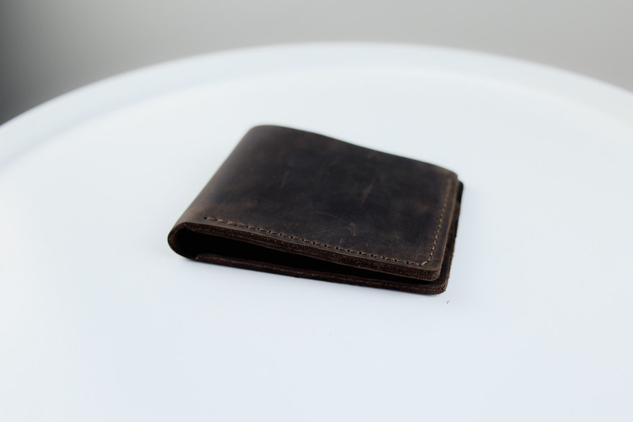 A dark brown leather bifold wallet with visible grain and contrast stitching, resting on a white circular surface.