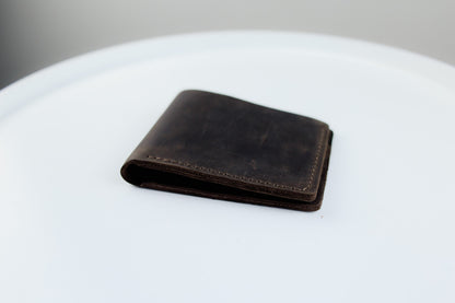 A dark brown leather bifold wallet with visible grain and contrast stitching, resting on a white circular surface.