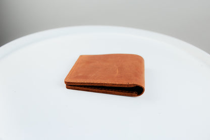 A brown leather bifold wallet with a distressed finish, folded and resting on a white circular surface against a neutral gray background.