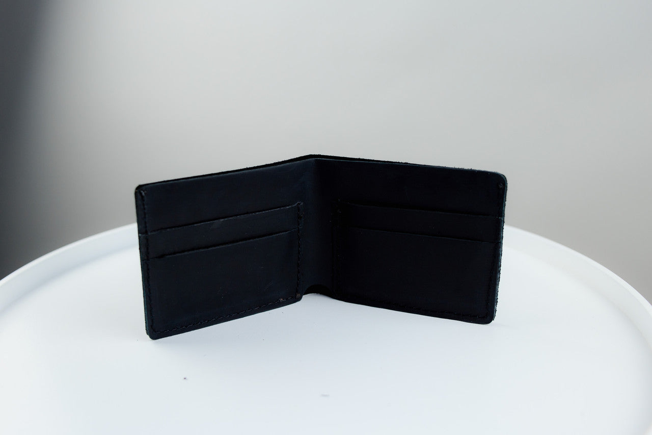 A black leather bifold wallet shown open, displaying its interior card slots, set on a white round surface against a neutral gray background.