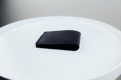 A black leather bifold wallet resting on a white circular platform against a neutral gray background.