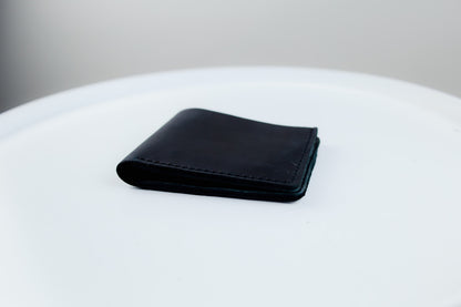 A black leather bifold wallet with visible stitching, resting on a white circular surface against a neutral gray background.