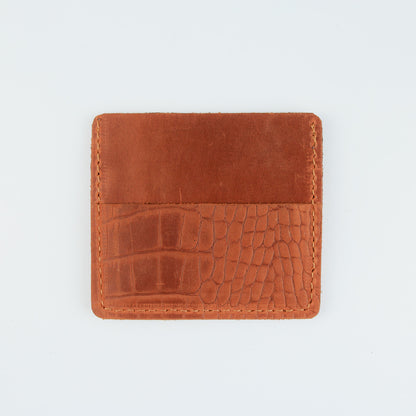 A square brown leather card holder or wallet with a split design, featuring a smooth leather top half and an embossed crocodile skin pattern on the bottom half, shown on a plain white background.