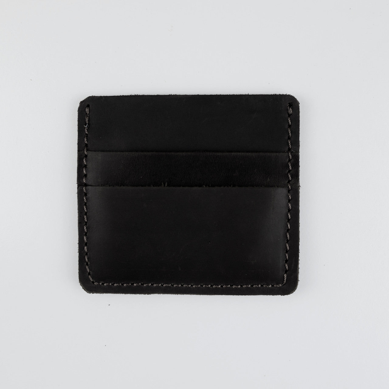 A minimalist black leather card holder with visible contrast stitching, photographed flat against a light gray background.