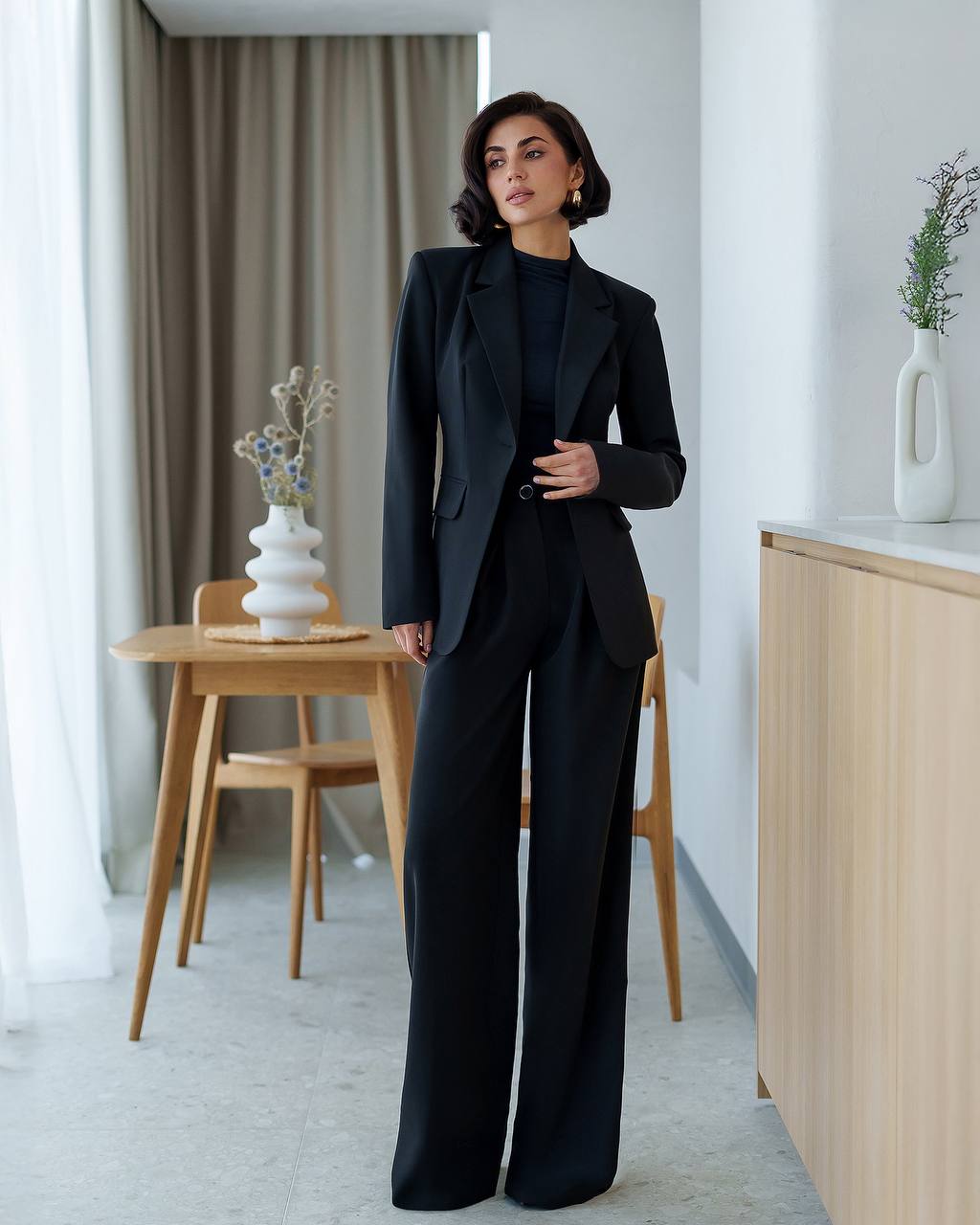 Woman in a black suit standing in a modern interior setting with a table and decorative items.