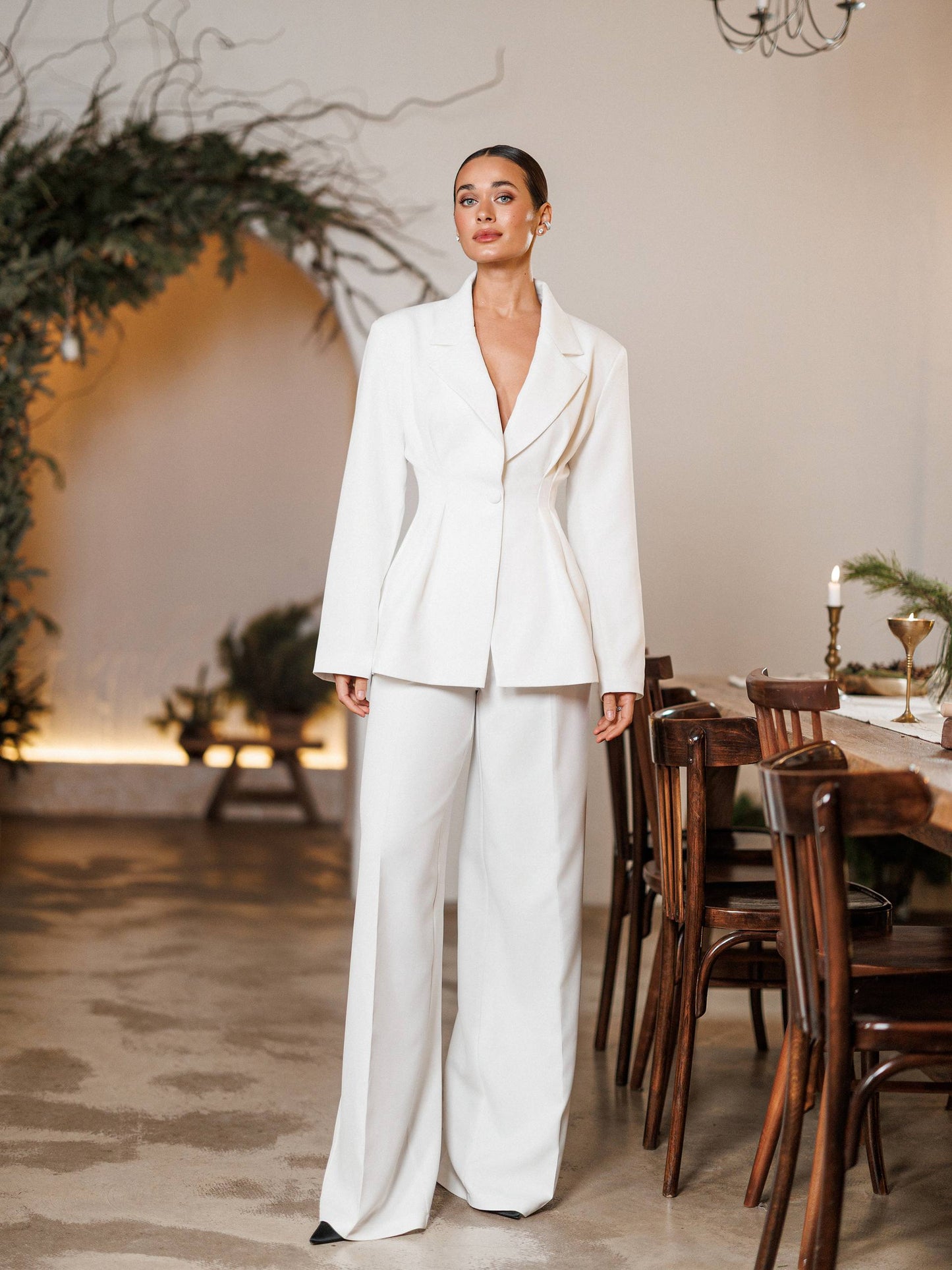 Woman in a white suit standing in an elegant indoor setting with wooden chairs and decorative plants.