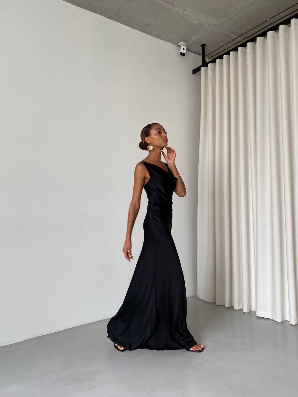 Woman in a black evening gown standing in a minimalistic room with white walls and curtains.