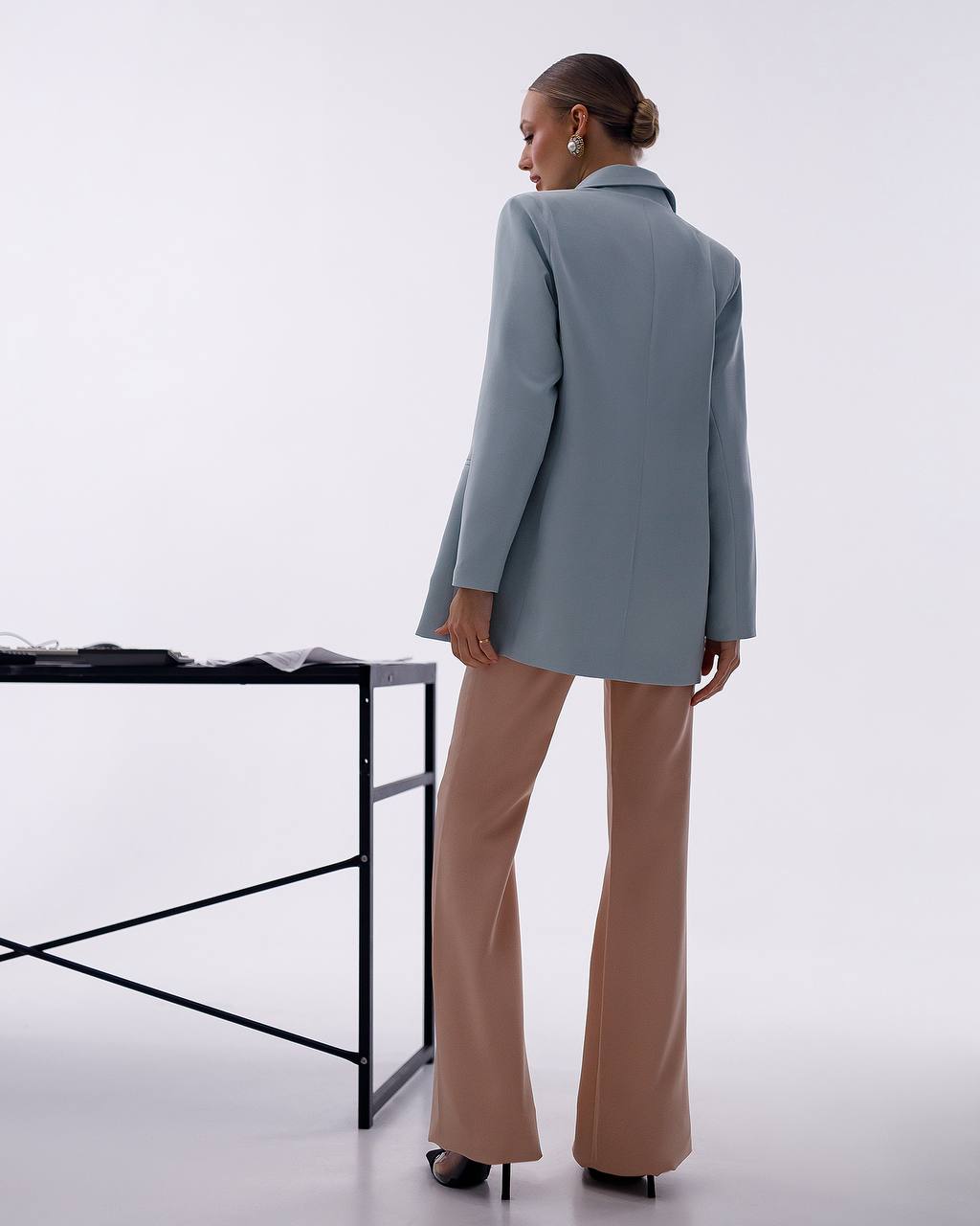 A model is shown from behind wearing a light blue blazer and beige flared trousers, standing next to a minimalist black metal table against a white studio background.