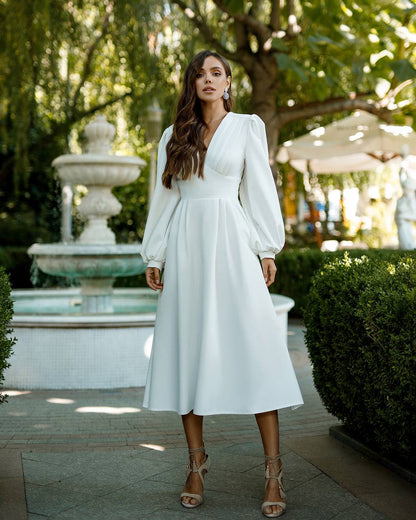 Woman in a white dress standing outdoors with a fountain and greenery in the background