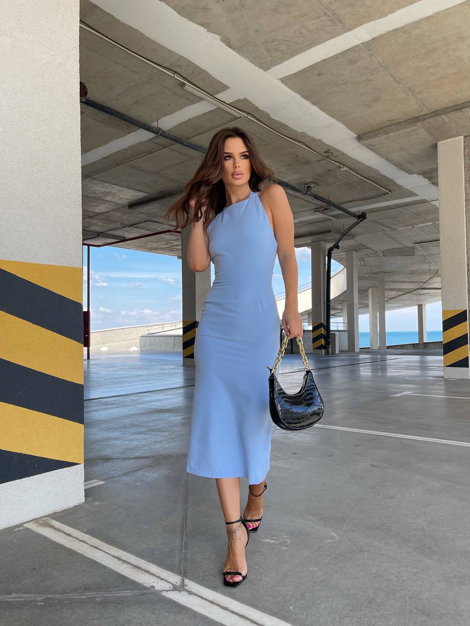 Woman in a light blue dress holding a black handbag in an underground parking garage.