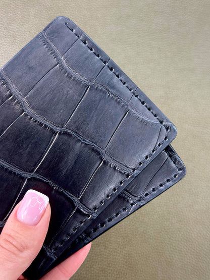 A close-up of a black crocodile-embossed leather wallet being held by a hand with light pink manicured nails, set against a textured olive green background.