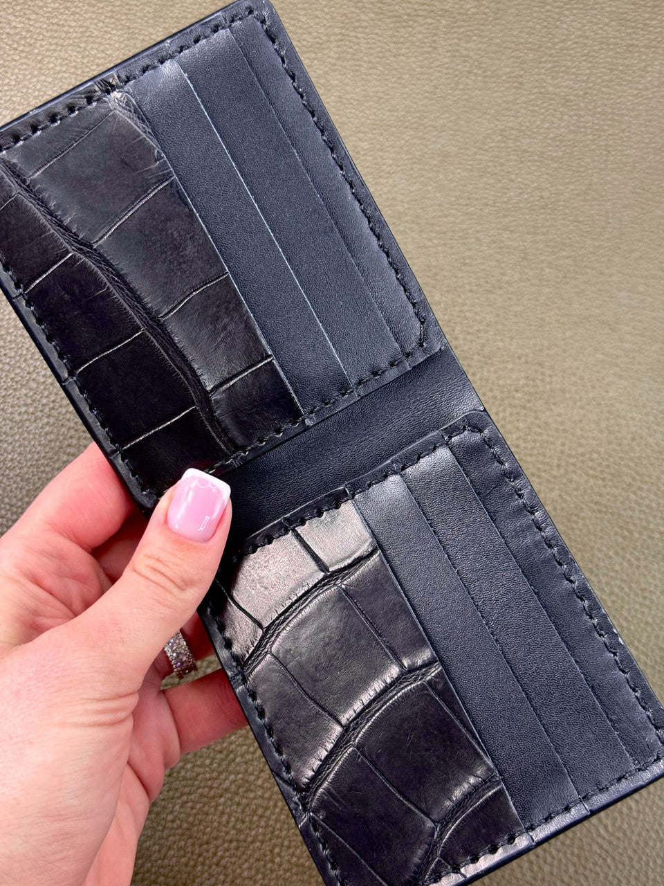 A close-up of a black leather bifold wallet with an alligator or crocodile-embossed texture, held open by a hand with pink nail polish against a textured beige background.