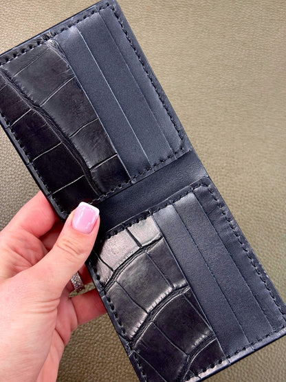 A close-up of a black leather bifold wallet with an alligator or crocodile-embossed texture, held open by a hand with pink nail polish against a textured beige background.
