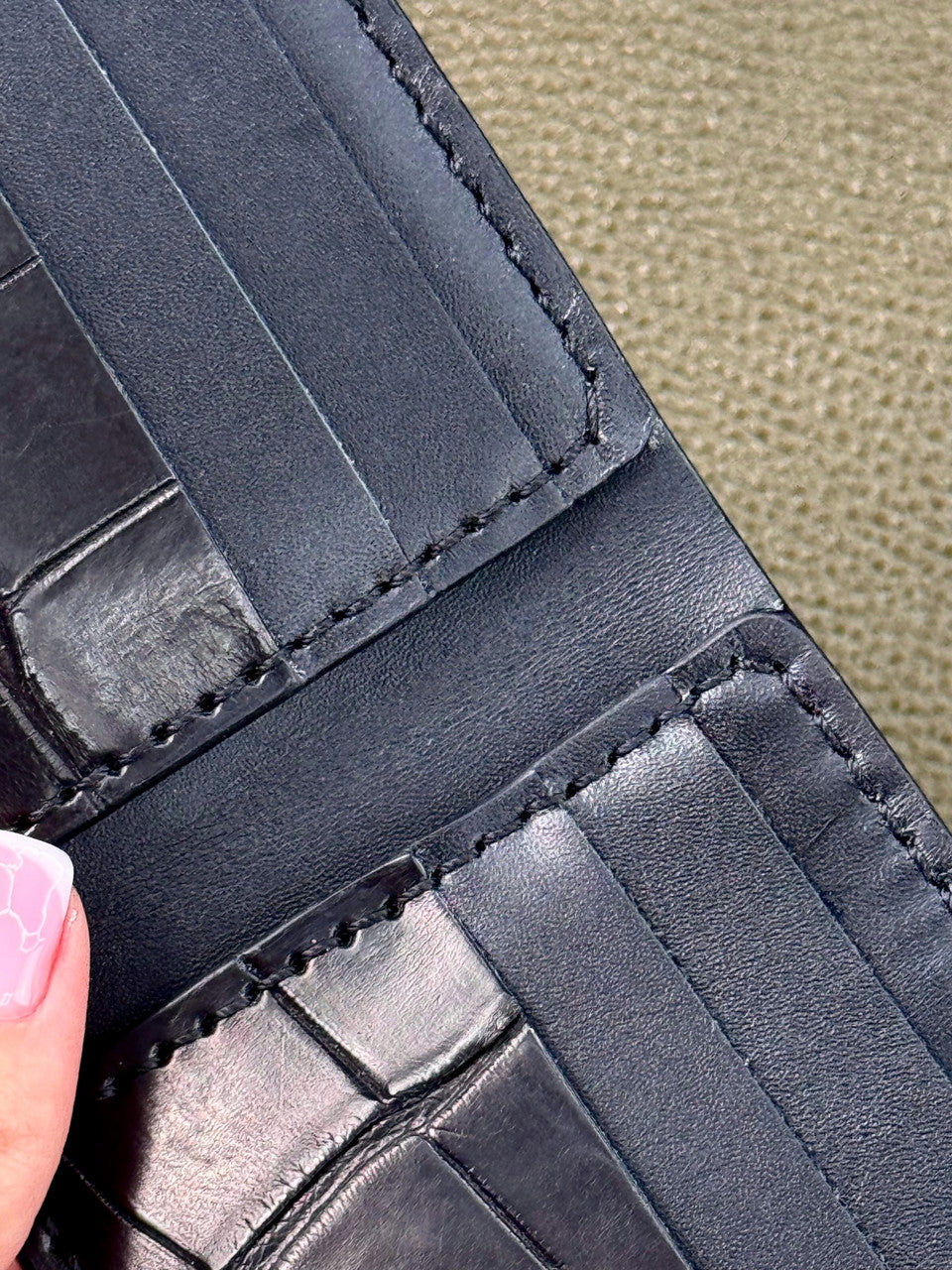Close-up of a black leather wallet showing interior card slots, stitching, and textured paneling, with a finger holding it open against a beige textured background.