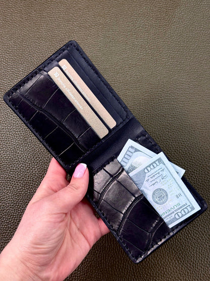 A hand holding open a black crocodile-embossed bifold wallet with card slots, showing a few cards and a $100 bill inside, set against a textured brown leather background.