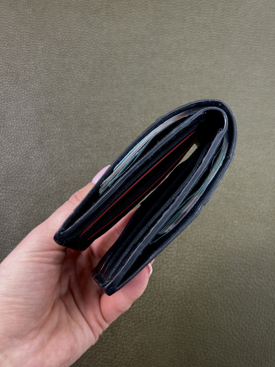 A black leather bifold wallet held in a hand, showing its interior card slots and thin red accent lines, against a textured olive green background.
