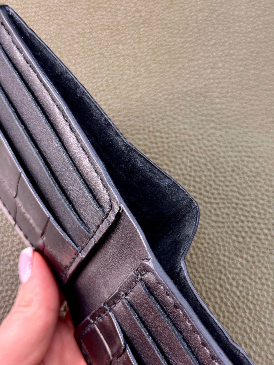 A close-up of a dark brown leather wallet being held by a hand, showing the interior card slots, stitching, and dark lining against a textured beige background.