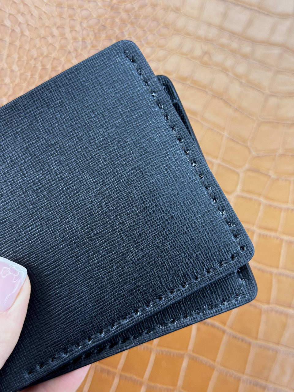A close-up of a black textured cardholder wallet being held by a hand, showing fine stitching and a tan crocodile-embossed background.